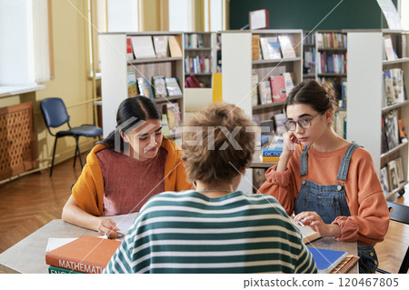 Two young female students sitting at a library table with books, engaging in studying and collaborating on their assignments. Bookshelves with a variety of books in the background 120467805