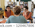 Group of students sitting at table discussing study materials and sharing ideas in library during study session, with books and shelves in background 120467806
