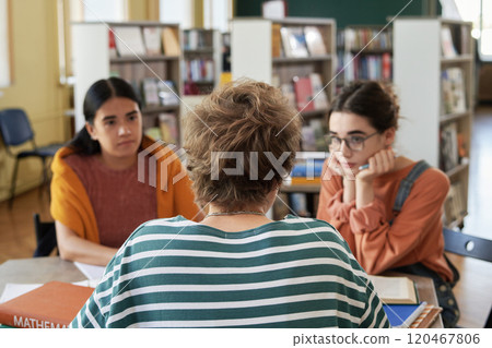 Group of students sitting at table discussing study materials and sharing ideas in library during study session, with books and shelves in background 120467806