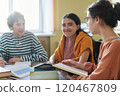 Group of friends collaborating on study session in library, sitting around table with books and notebooks, engaged in discussion and smiling 120467809