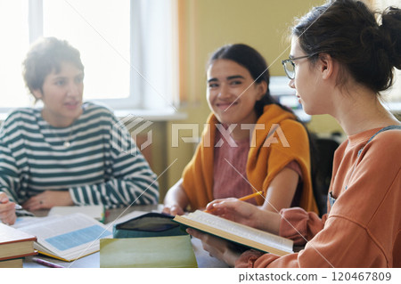 Group of friends collaborating on study session in library, sitting around table with books and notebooks, engaged in discussion and smiling 120467809