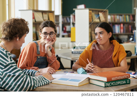 Three students sitting in library discussing class project while surrounded by books and educational materials where one person smiling warmly and the others listening attentively 120467811