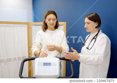 A female cardiologist tests a patient during a bike workout to check the cardiovascular system in the clinic's medical office. A young woman trains on an exercise bike. A female cardiologist tests a patient during a bike workout to check the cardiovascular system in the clinic's medical office. A young woman trains on an exercise bike. 120467875
