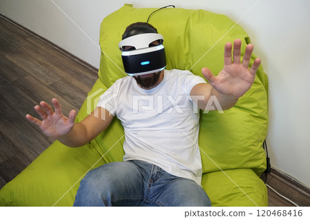 Man seated on green bean bag, wearing VR headset, extending his hands in virtual environment Man seated on green bean bag, wearing VR headset, extending his hands in virtual environment 120468416