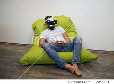 Man is seated on green bean bag, using VR headset and holding controller Man is seated on green bean bag, using VR headset and holding controller 120468418