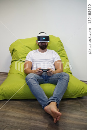 Man wearing VR headset, sitting comfortably on green bean bag while gaming with controller 120468420