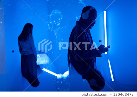 Two individuals immersed in virtual reality experience in a dark room illuminated by blue neon lights. Man holding device in foreground while woman facing screen background Two individuals immersed in virtual reality experience in a dark room illuminated by blue neon lights. Man holding device in foreground while woman facing screen background 120468472