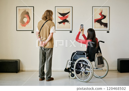 Two people in modern gallery observing and photographing abstract art pieces displayed on wall. One person is standing while other is sitting in a wheelchair 120468502