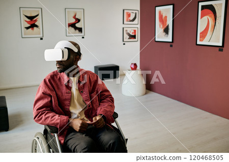 Man in wheelchair exploring modern art gallery using virtual reality headset with abstract paintings on walls and contemporary decor elements surrounding him 120468505