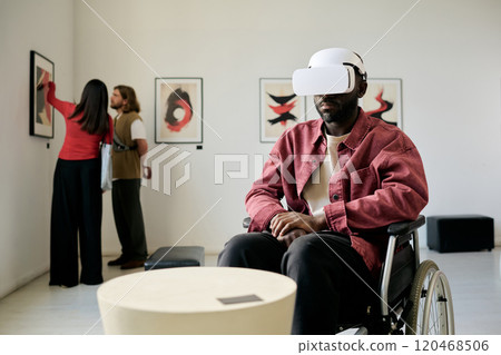 Man wearing VR headset sitting in wheelchair in art gallery, immersed in virtual experience while visitors in background examine paintings on wall 120468506
