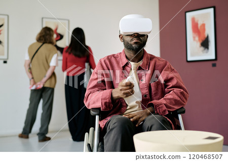 Man in wheelchair wearing VR headset, engaging with digital art in contemporary gallery space, while others observe paintings on walls. Immersive technology enhancing art experience 120468507