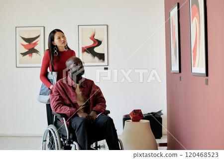 Woman standing behind wheelchair, looking at contemporary artworks in gallery. Man in wheelchair studying paintings on wall with focus and interest Woman standing behind wheelchair, looking at contemporary artworks in gallery. Man in wheelchair studying paintings on wall with focus and interest 120468523