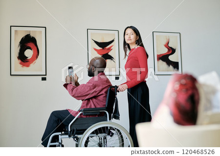 Two people enjoying vibrant abstract art pieces in modern gallery, one being in wheelchair with the other standing nearby while holding another artwork 120468526