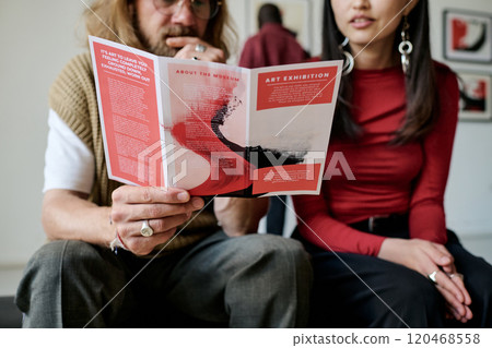 Two people sitting closely together discussing details of an art exhibition pamphlet, with additional background figures visible. Casual yet focused atmosphere shared by individuals 120468558