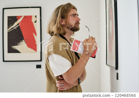 Man with glasses and notebook viewing art in contemporary gallery space, absorbed in thought, appearing deeply engaged with artwork Man with glasses and notebook viewing art in contemporary gallery space, absorbed in thought, appearing deeply engaged with artwork 120468568