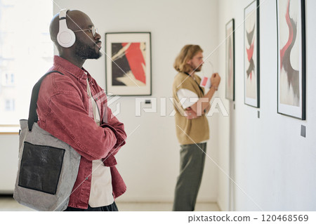 African American man wearing headphones examining modern paintings in art gallery. Another person in background looking at artwork, creating immersive viewing experience African American man wearing headphones examining modern paintings in art gallery. Another person in background looking at artwork, creating immersive viewing experience 120468569
