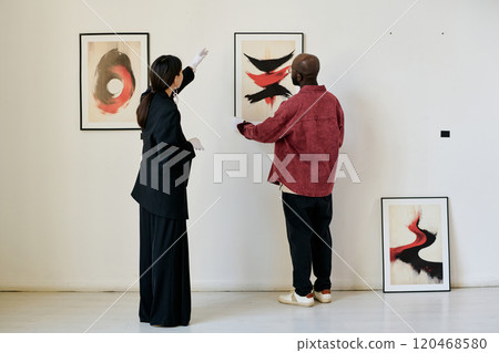 Two people admiring and discussing framed abstract paintings on a white gallery wall. The artworks feature bold colors and dynamic forms creating a striking visual experience 120468580