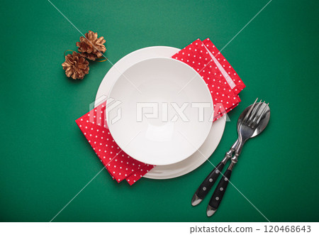 Christmas table setting with empty plate, golden pine cones, and red napkin on green background 120468643