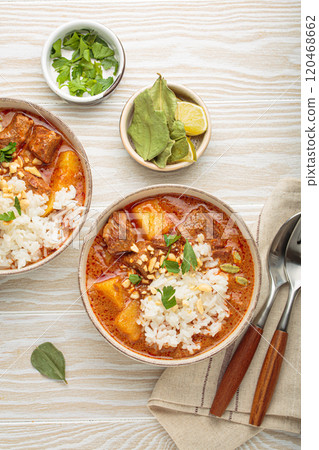 Two bowls of delicious massaman curry with beef, potatoes, rice, and peanuts, garnished with parsley on white wooden background, Thai cuisine 120468662
