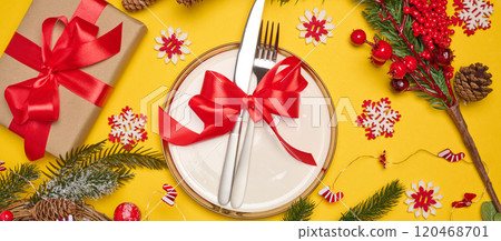 Festive composition with a plate and cutlery tied with a red ribbon, a gift, fir branches, pine cones and decorative snowflakes on a bright yellow background Festive composition with a plate and cutlery tied with a red ribbon, a gift, fir branches, pine cones and decorative snowflakes on a bright yellow background 120468701