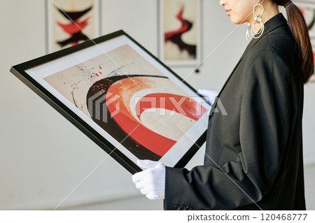 Person holding framed abstract painting in modern art gallery while examining details carefully. Exhibits and white wall in background with soft focus creating a serene atmosphere 120468777