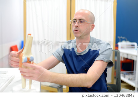 Orthopedist shows the patient an anatomical model of knee joint during appointment in modern clinic. Doctor explains features of structure. Rheumatologist tells about problems of treating disease. 120468842