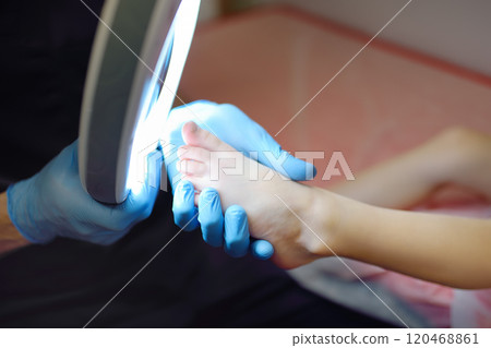 A caring doctor checks toenails of a small child using lamp with magnifying glass. A dermatologist looks at a rash on the toe of a girl A caring doctor checks toenails of a small child using lamp with magnifying glass. A dermatologist looks at a rash on the toe of a girl 120468861