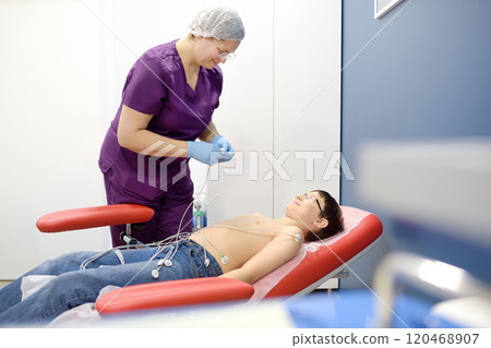 Child under ECG test with doctor. Boy patient lying in medical bed with vacuum sensors. A nurse performs an electrocardiogram of the heart in office of a medical clinic. Child under ECG test with doctor. Boy patient lying in medical bed with vacuum sensors. A nurse performs an electrocardiogram of the heart in office of a medical clinic. 120468907