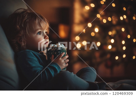 Young toddler is sitting by the fireplace, holding a mug of hot chocolate with marshmallows, enjoying the warm Christmas atmosphere 120469309