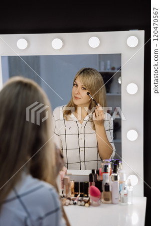 Woman applying makeup in front of a vanity mirror with bright lights for a flawless look 120470567