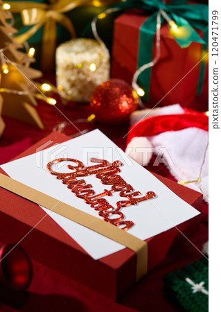 A white card with a sparkling Merry Christmas wish on a red gift box, surrounded by various Christmas decorations and yellow LED lights. High angle view, space for design A white card with a sparkling Merry Christmas wish on a red gift box, surrounded by various Christmas decorations and yellow LED lights. High angle view, space for design 120471799