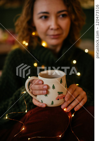 Mid-aged woman sitting indoors, holding a Christmas-themed mug and surrounded by warm holiday lights, creating a cozy and serene Christmas atmosphere Mid-aged woman sitting indoors, holding a Christmas-themed mug and surrounded by warm holiday lights, creating a cozy and serene Christmas atmosphere 120473516