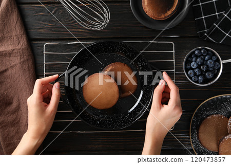Chocolate pancakes on plates and frying pan on grid, hands, bowl with berries on wooden background, top view Chocolate pancakes on plates and frying pan on grid, hands, bowl with berries on wooden background, top view 120473857