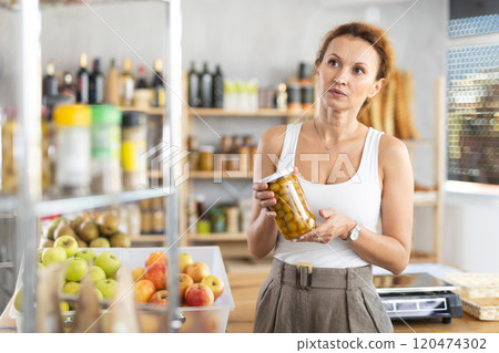 Female shopper carefully choosing jar of canned olives in grocery supermarket Female shopper carefully choosing jar of canned olives in grocery supermarket 120474302