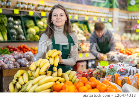 Young female seller holding bananas standing by counter in grocery market Young female seller holding bananas standing by counter in grocery market 120474318
