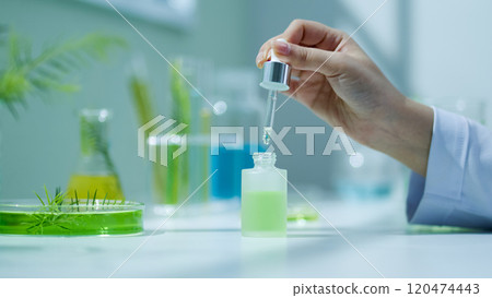 A scientist holding a dropper cap of a white opaque serum bottle containing a tea tree essence, surrounded by fully equipped lab equipment, a front view of the photo in the lab backdrop. 120474443