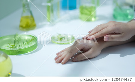 A front view of a researcher hands applying cream with tea tree as the main ingredient to testing texture. Modern labs feature a tabletop surface fully equipped lab supplies. A front view of a researcher hands applying cream with tea tree as the main ingredient to testing texture. Modern labs feature a tabletop surface fully equipped lab supplies. 120474487