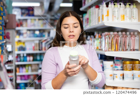 Young brunette holding hair care lotion and exploring it in beauty store 120474496