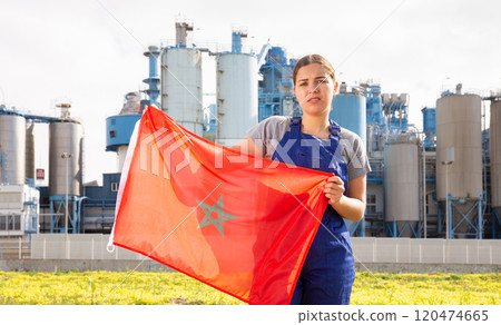 upset woman in blue worker s overalls stands near country factory and holds Moroccan flag 120474665