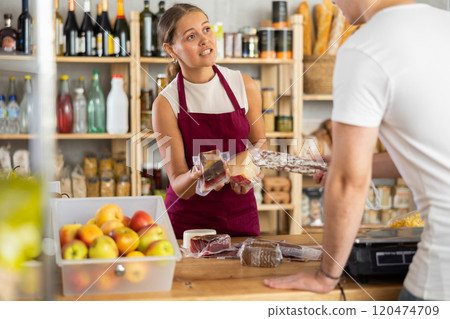 Friendly female salesperson offers to buy fresh cheese and dried meat to a male customer at grocery store 120474709