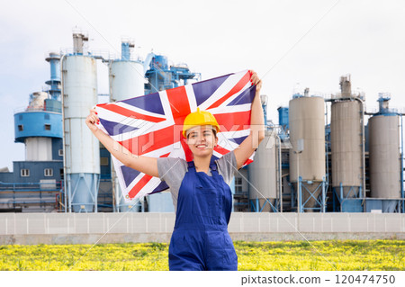 Positive young female worker in helmet waving national flag of Great Britain while standing in front of big tanks at refinery factory on sunny summer day 120474750