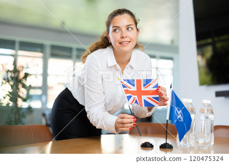 Woman secretary prepares an office for negotiations - she places flags of NATO (OTAN) and Great Britain table Woman secretary prepares an office for negotiations - she places flags of NATO (OTAN) and Great Britain table 120475324