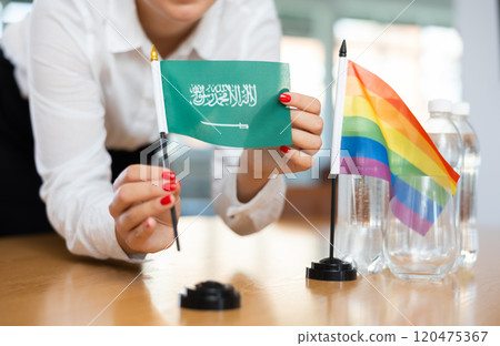 Young woman putting Saudi Arabian and LGBT flags on table in office 120475367