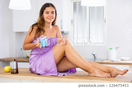 Portrait of young woman sitting on table in kitchen and drinking coffee 120475451