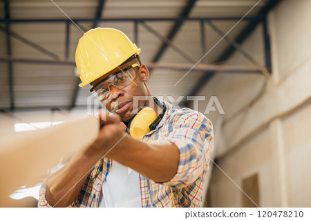 African American carpenter inspecting a piece of wood while carrying a wooden plank in a workshop. construction work, wood quality inspection, and carpentry craftsmanship, National Carpenters Day 120478210
