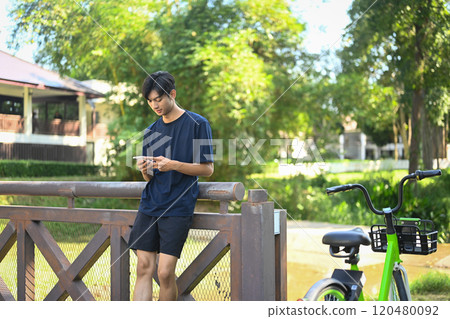 Young asian man using mobile phone, leaning on a bridge next to a bicycle in a scenic park Young asian man using mobile phone, leaning on a bridge next to a bicycle in a scenic park 120480092