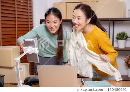 Two young asian woman live online clothing store while showing clothing with tablet and laptop. 120480691