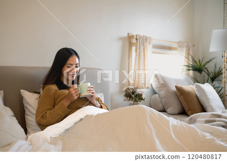 Pleased young woman enjoying warm drink sitting under a white duvet in bright bedroom Pleased young woman enjoying warm drink sitting under a white duvet in bright bedroom 120480817
