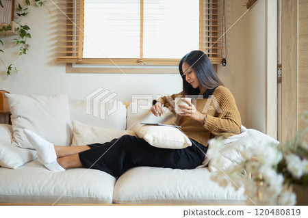 Beautiful young woman holding a mug and using digital tablet on soft sofa Beautiful young woman holding a mug and using digital tablet on soft sofa 120480819