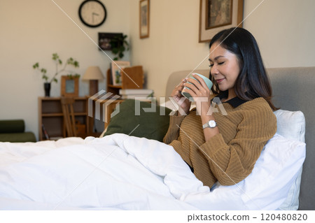 Pleased young woman enjoying warm drink sitting under a white duvet in bright bedroom Pleased young woman enjoying warm drink sitting under a white duvet in bright bedroom 120480820